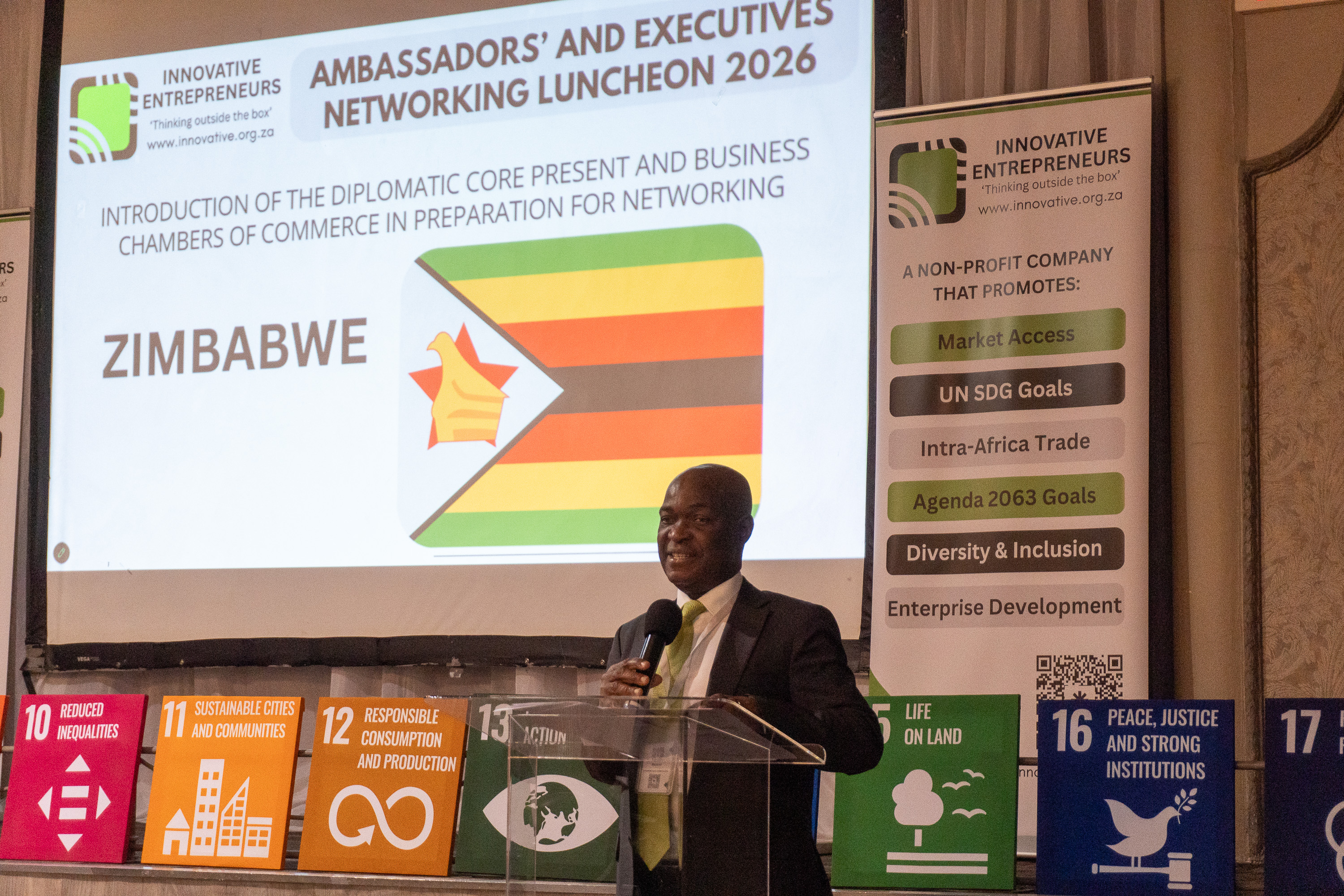 A Zimbabwean diplomatic representative addresses AENL 2026 with the Zimbabwe flag projected behind
