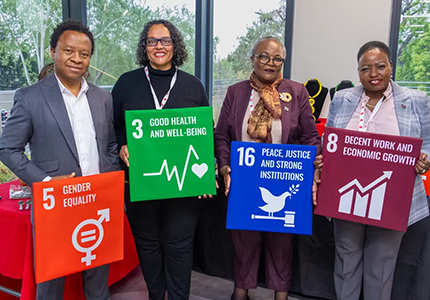 Four individuals, two men and two women, are standing and smiling while holding large square signs representing United Nations Sustainable Development Goals. From left to right: a man in a grey suit holds '5 Gender Equality,' a woman in black holds '3 Good Health and Well-being,' another woman in a patterned scarf holds '16 Peace, Justice and Strong Institutions,' and a woman in a grey suit holds '8 Decent Work and Economic Growth.' They appear to be at an event or conference, demonstrating commitment to global goals. Keywords: entrepreneurship and SDGs, UN SDG-aligned business models, DEI in business South Africa, empowering women in business, economic inclusion initiatives, inclusive economy summit.