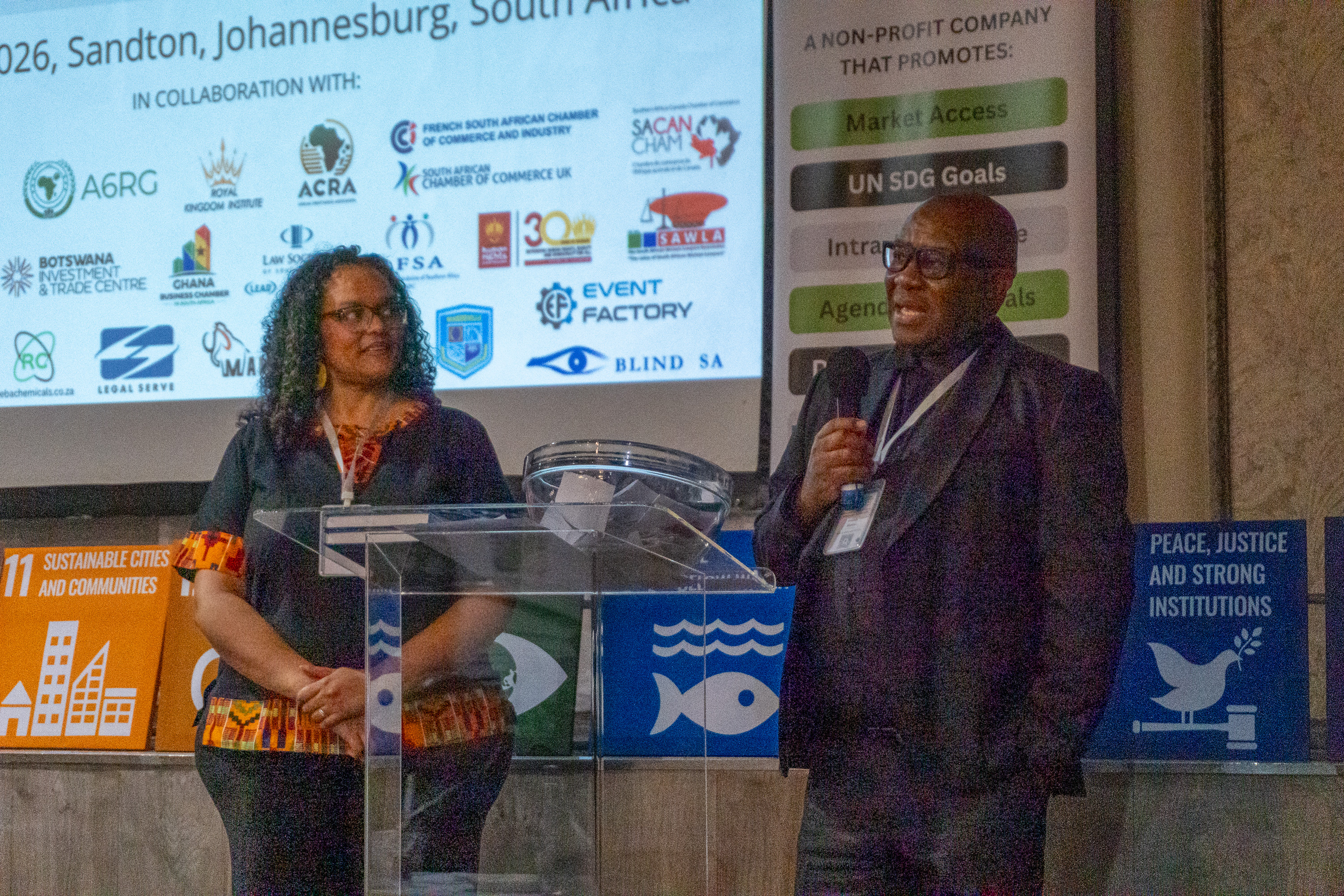 Two speakers address the audience at AENL 2026 with the full sponsor and partner logo wall projected behind