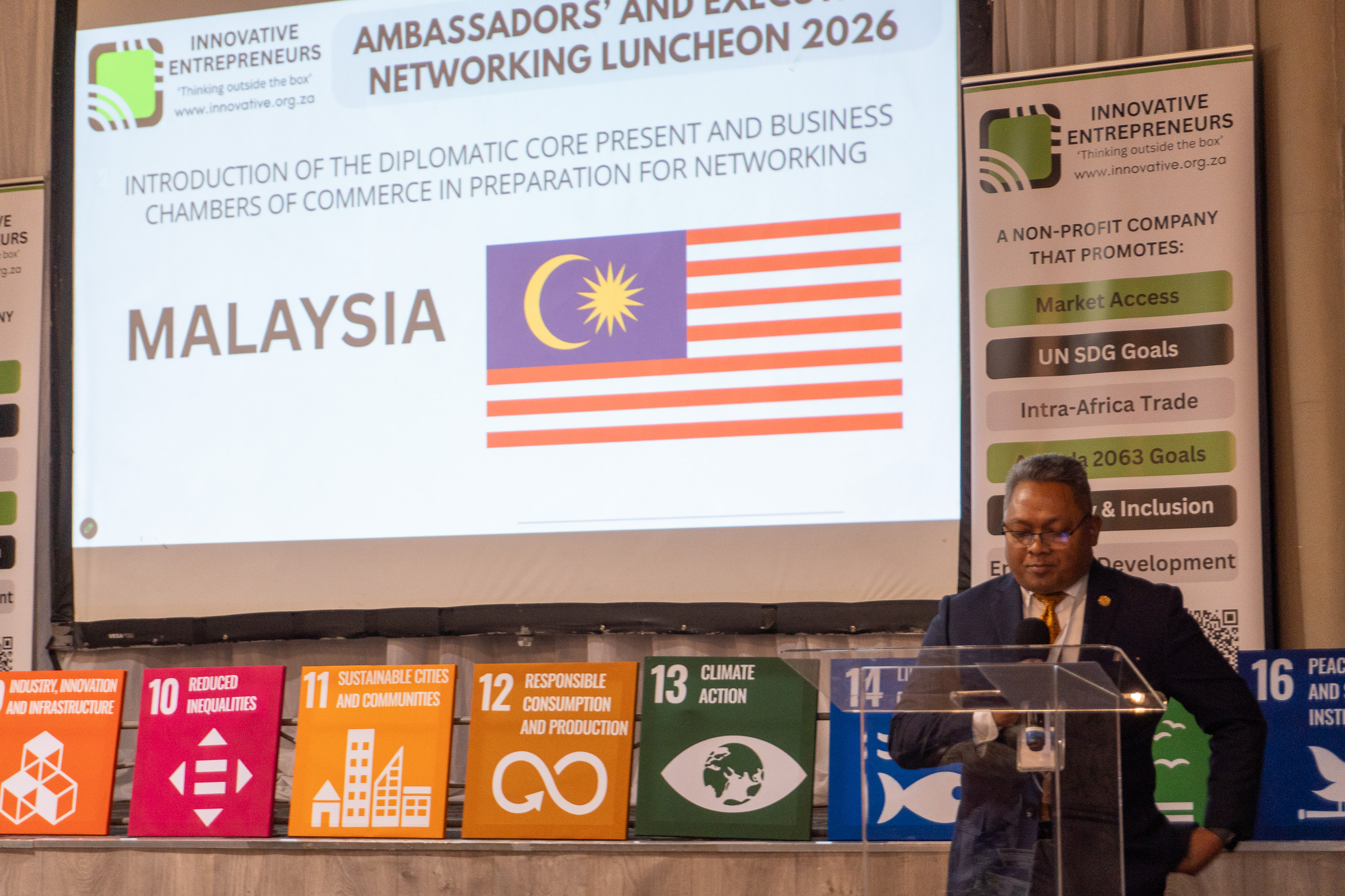 A Malaysian diplomatic representative addresses AENL 2026 with the Malaysia flag projected behind